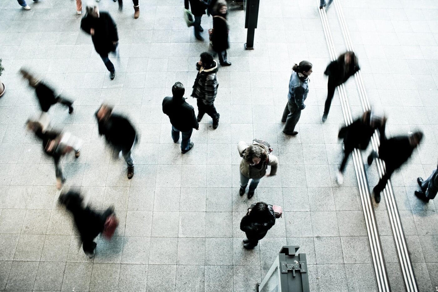 A large group of people walking through a crowded urban street, carrying backpacks, bags, and personal belongings as they attempt to leave the city on foot. The scene is filled with tension, with individuals navigating past stalled vehicles and closed shops under the shadow of tall buildings. The image highlights the challenges of evacuation in densely populated areas, where mass transportation and resources like fuel and food may be unavailable. This moment underscores the importance of having an emergency plan, situational awareness, and preparedness strategies, including bug-out bags and alternative routes, for navigating chaotic urban environments during disasters.