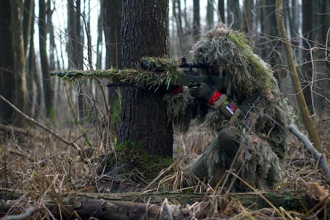 camouflaged hunter taking aim behind a tree.